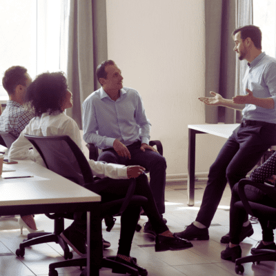 A man leaning against a table explains something to three seated employees.