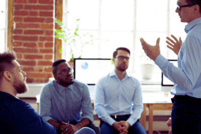 A man explaining and gesturing to three seated coworkers.