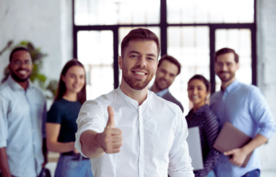 A smiling man holding a thumbs-up as his coworkers stand and smile behind him.